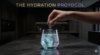 A cinematic, macro shot of a hand dropping a pinch of coarse, crystalline sea salt into a glass of pure, sparkling water. The water rests on a dark marble countertop and emits a subtle cyan glow. Dark luxury aesthetic. Integrated title: THE HYDRATION PROTOCOL.